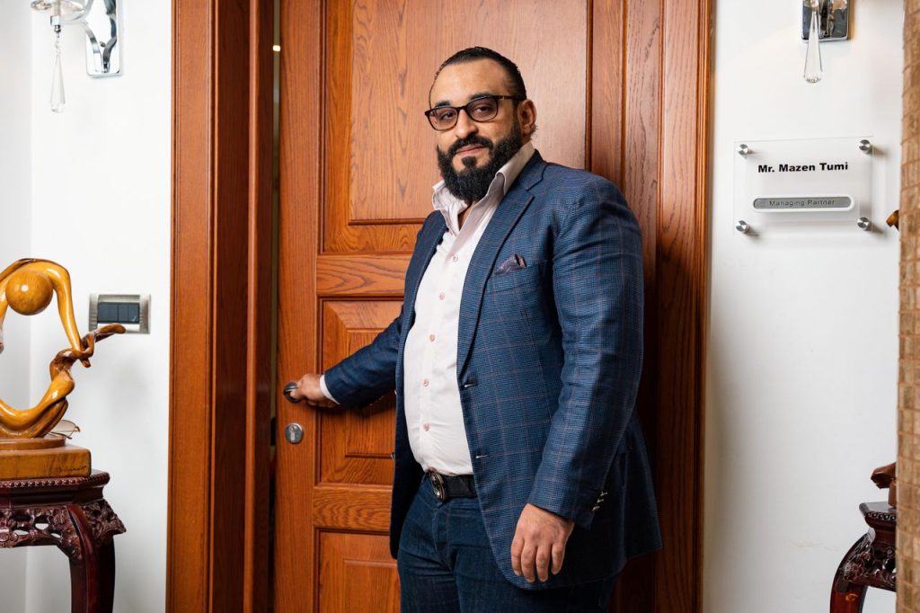 A bearded man in a blue suit and eyeglasses stands confidently by a wooden door inside a well-decorated office.