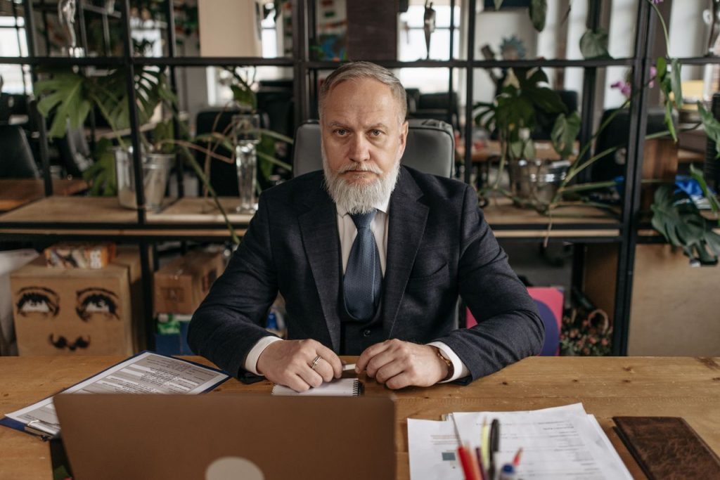 Elderly businessman in a suit at his office desk with papers and a laptop, serious expression.